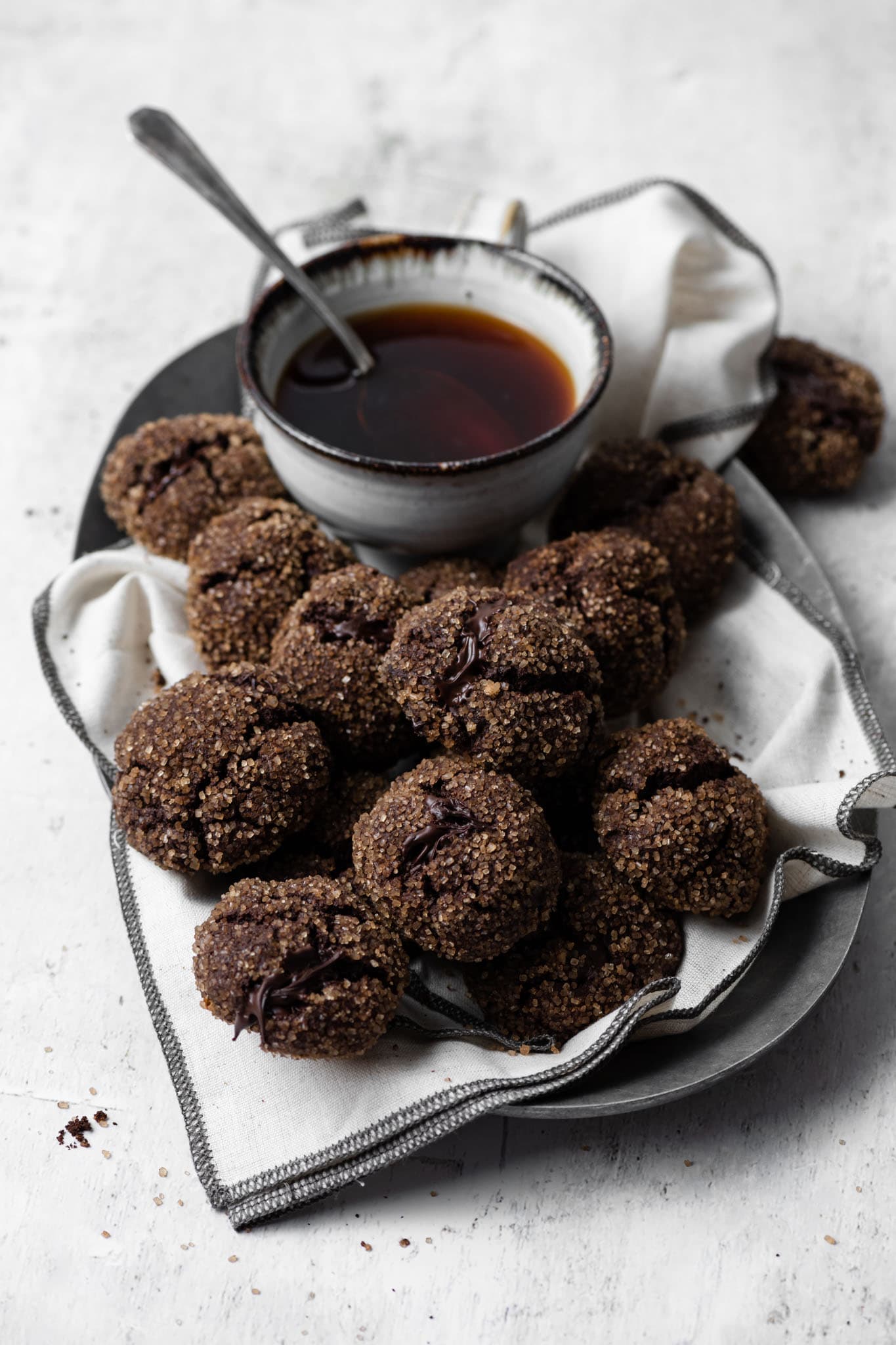 Oval metal serving plate full of round chocolate cookies rolled in sugar on a linen napkin with a cup of coffee.