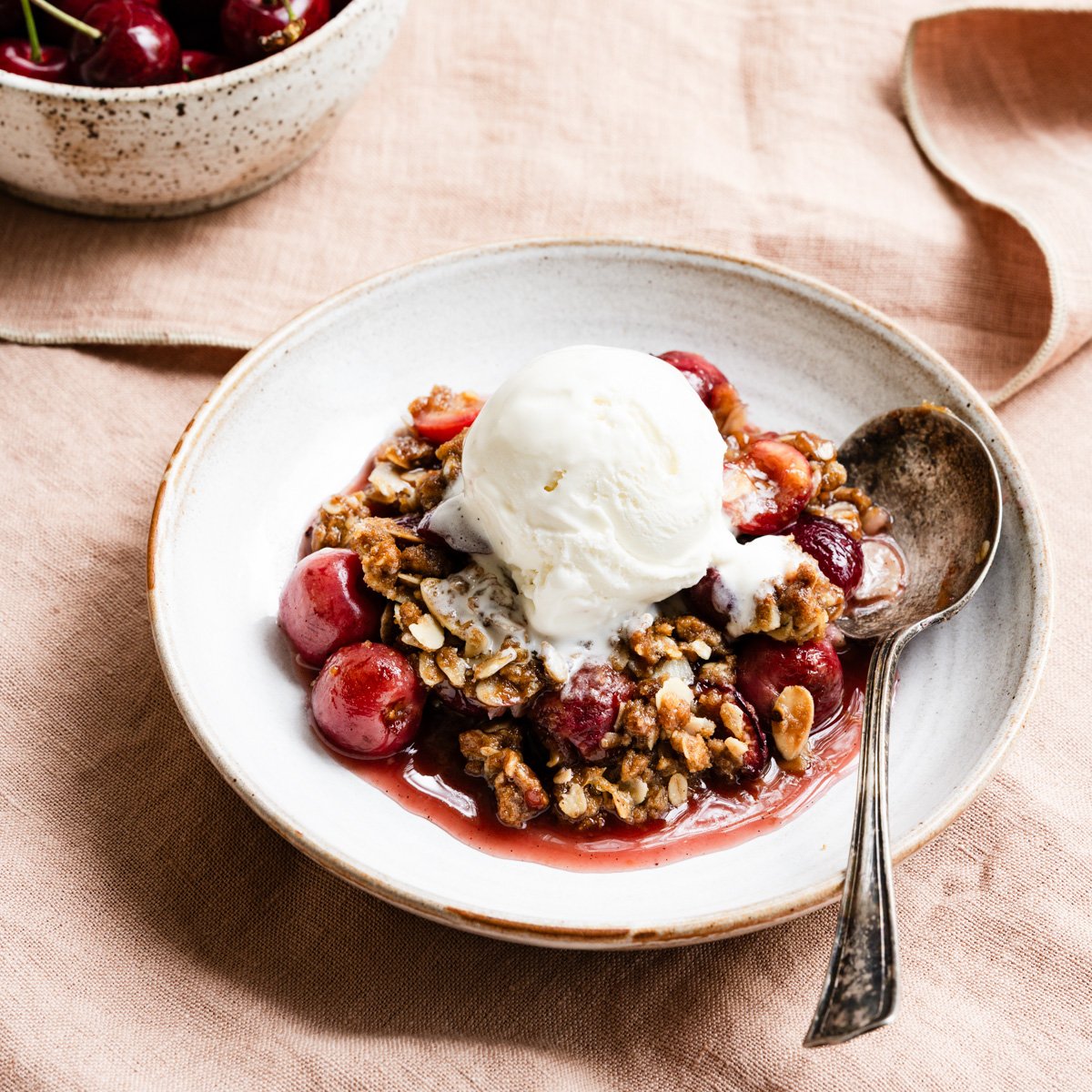 Bowl filled with cherry crisp and topped with a scoop of vanilla ice cream on a table cloth next to a bowl of cherries.