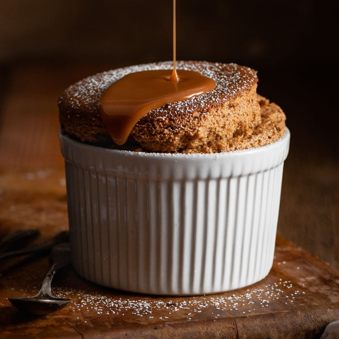 A cooked souffle in a ramekin with butterscotch sauce being poured over the top.