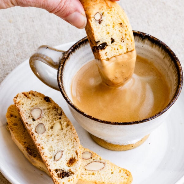 Hand dipping a biscotti cookie into a cup of coffee on a plate next to more biscotti.