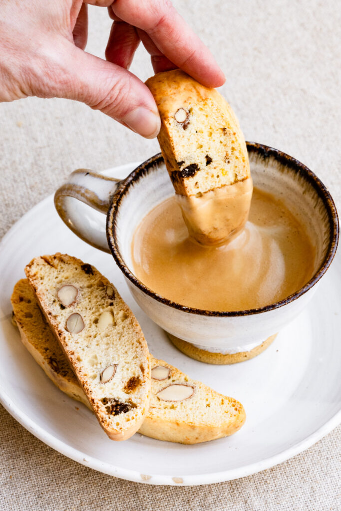 Hand dipping a biscotti cookie into a cup of coffee on a plate next to more biscotti.