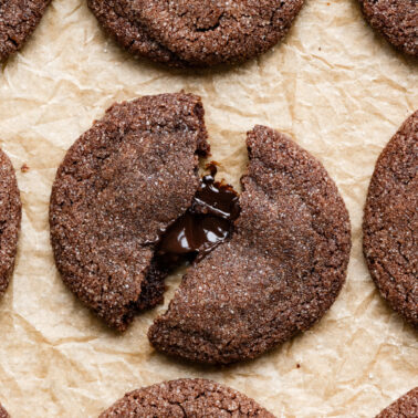 Overhead of chocolate cookies coated in sugar on baking tray, one is broken in half with chocolate filling.