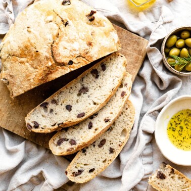 Overhead view of a loaf of olive bread on a wood cutting board with three slices cut. The board is on a linen covered table next to bowls of olives and olive oil.