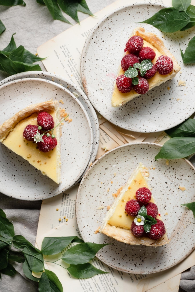 Three slices of lemon cheesecake topped with raspberries on three speckled plates.