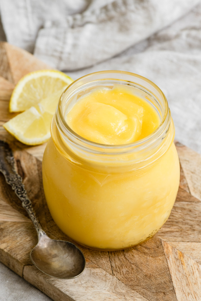 Glass jar of lemon curd on a wooden cutting board next to a spoon and sliced lemons.