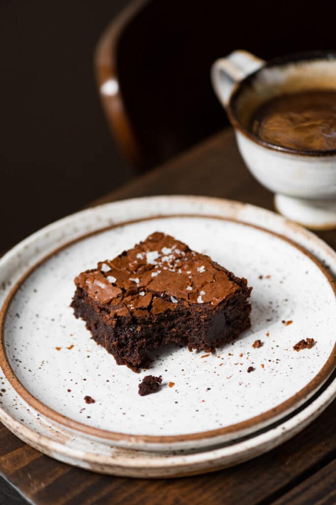 Chocolate brownie with bite removed on plate next to a cup of coffee. 
