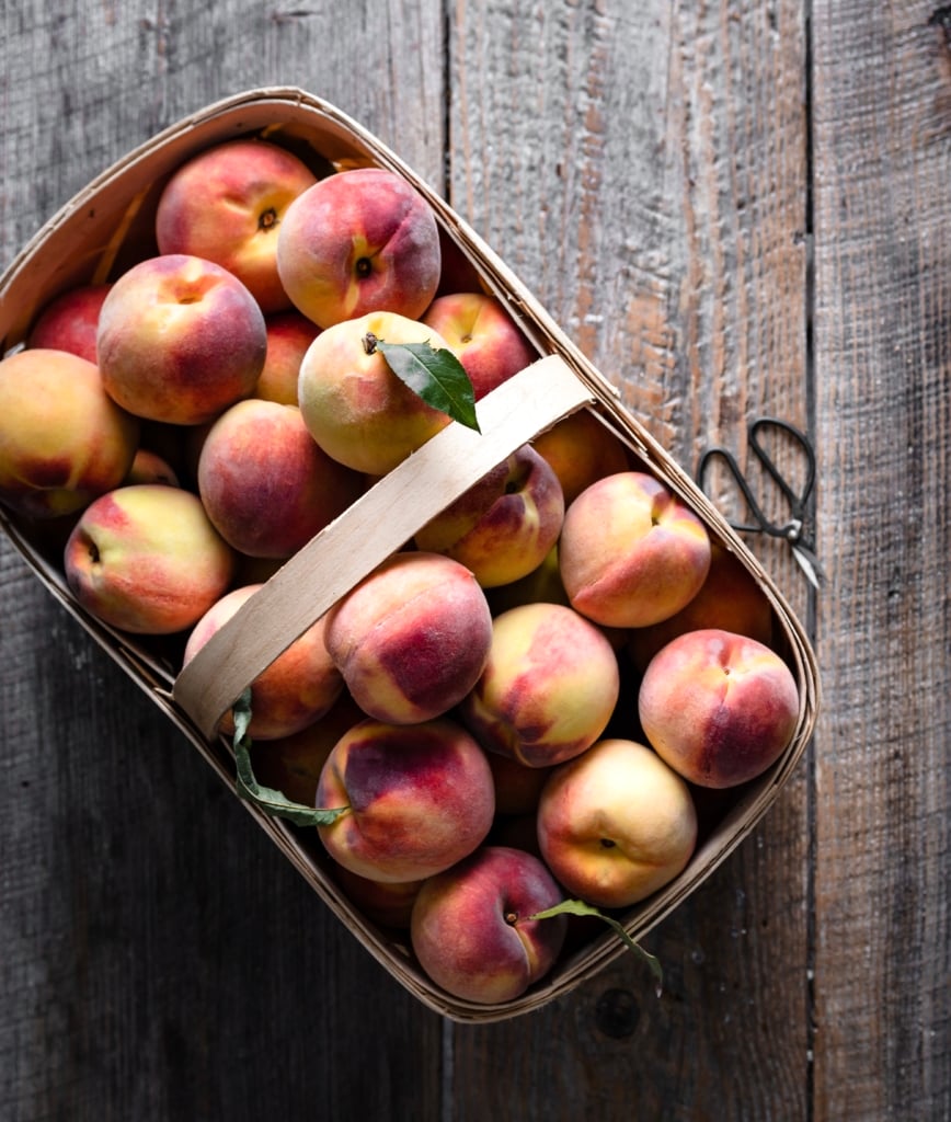 Overhead view of a basket filled with yellow peaches on a wooden table.