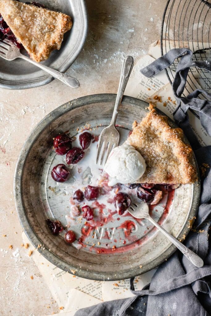 Slice of cherry pie topped with melted vanilla ice cream in a pie dish on table.