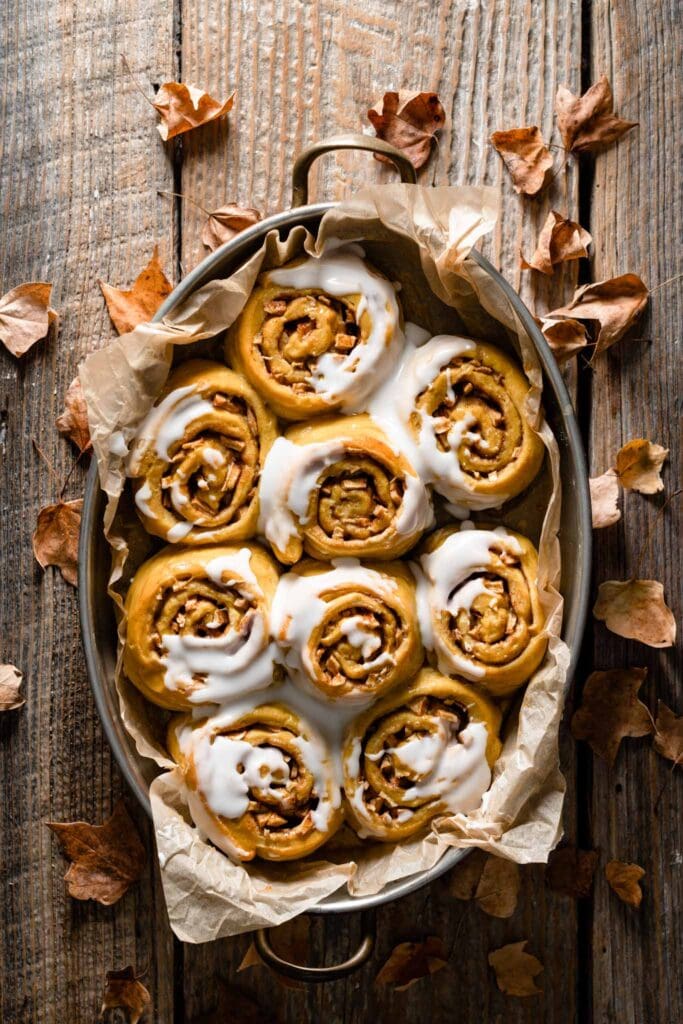Tray of baked pumpkin cinnamon rolls covered in frosting on table.