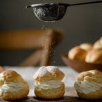 Cream puffs on a board being sprinkled with powdered sugar.