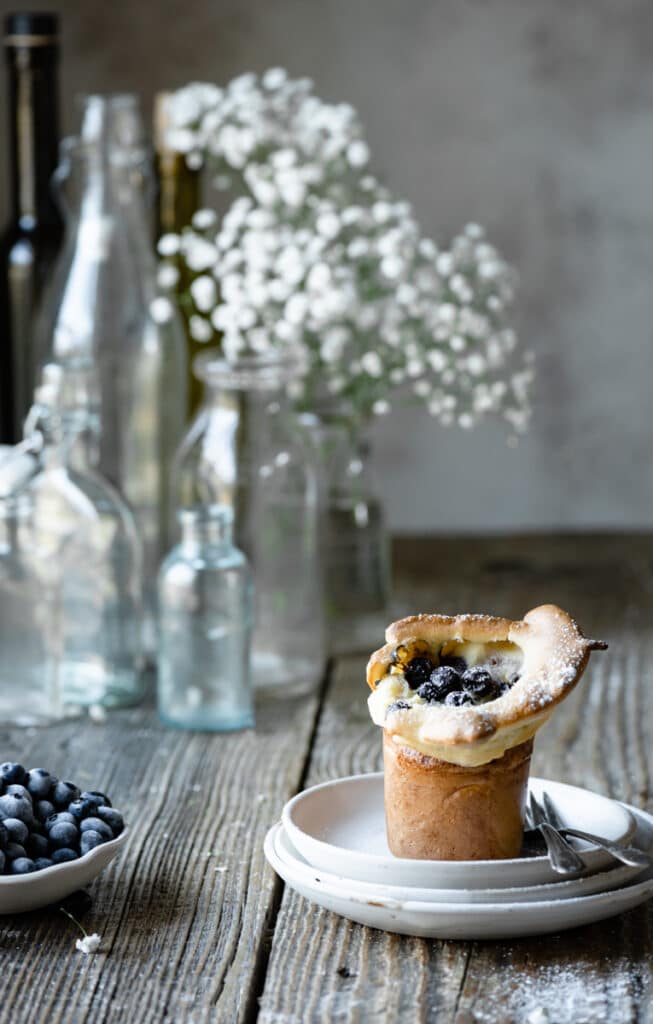 Single popover on a stack of plates on wood table next to flowers and a bowl of blueberries.