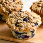 Three large blueberry crumble muffins on a wood cutting board.