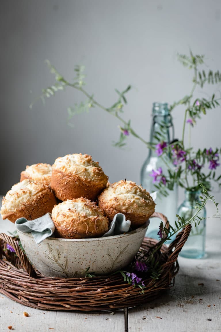 Bowl full of coconut topped muffins on a white table next to flowers.