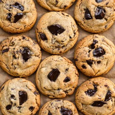 Overhead view of peanut butter cookies with dark chunks of chocolate aligned in rows on a table cloth.