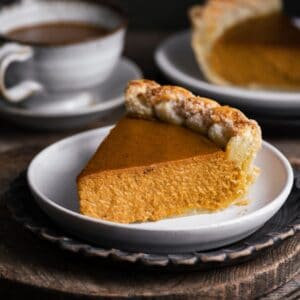 Close up of a slice of pumpkin pie on a white plate next to a cup of coffee.