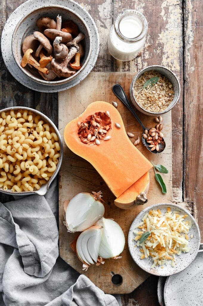 Sliced butternut squash, bowl of dry noodles and bowl of mushrooms next to shredded cheese on a wood cutting board.