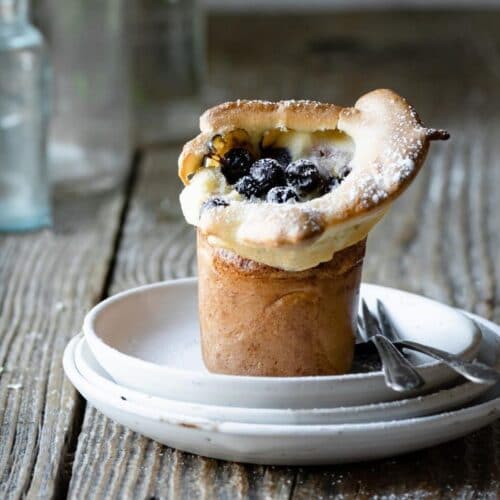 Closeup of a popover pastry on stack of white plates on table, topped with blueberries.