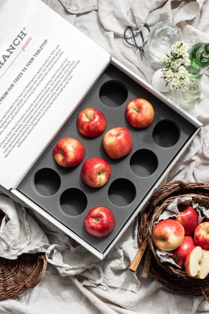 Open large box containing red apples on a linen cloth next to a basket of red apples.