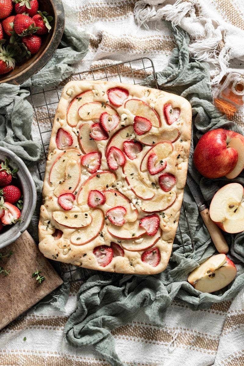 Fruit topped focaccia bread on table next to apples and sliced strawberries.