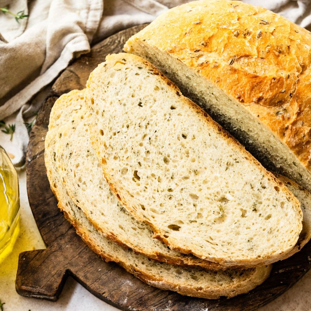 Round loaf of bread sliced on a wooden cutting board next to glass jar of olive oil.