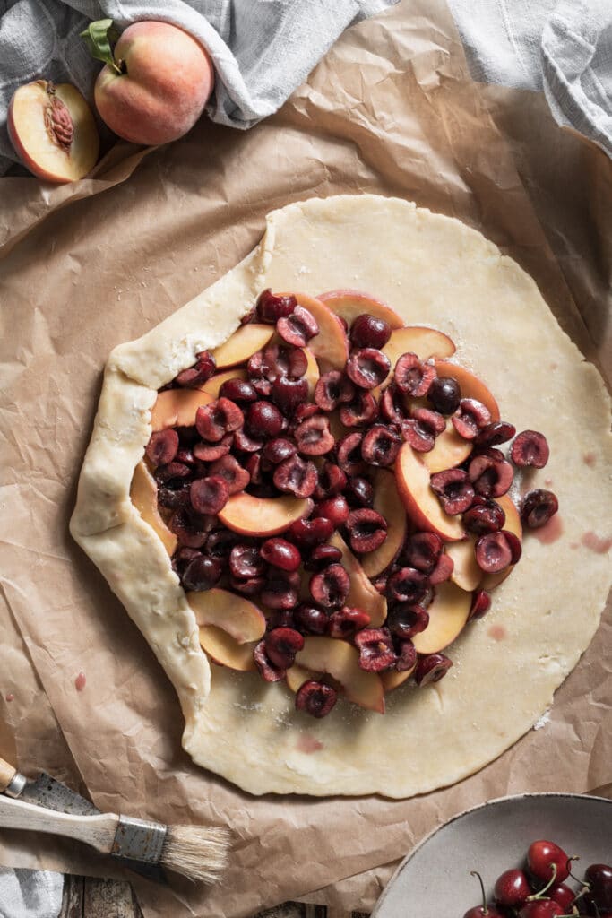 Unbaked pie crust on parchment paper filled with sliced cherries and peaches.
