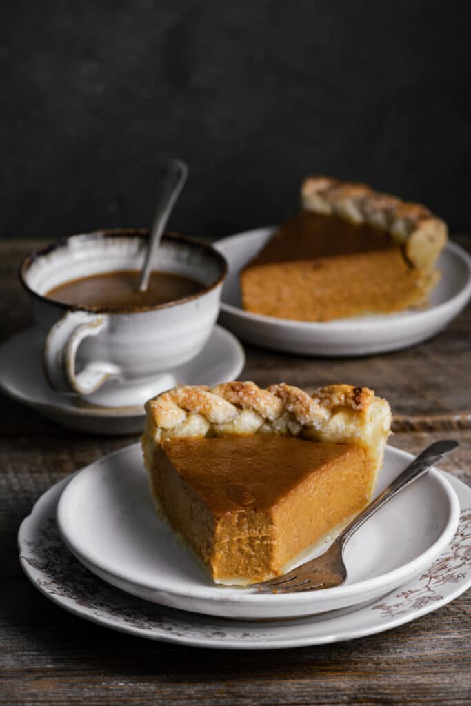 Two slices of pumpkin pie on white plates next to a cup of coffee on wood table.