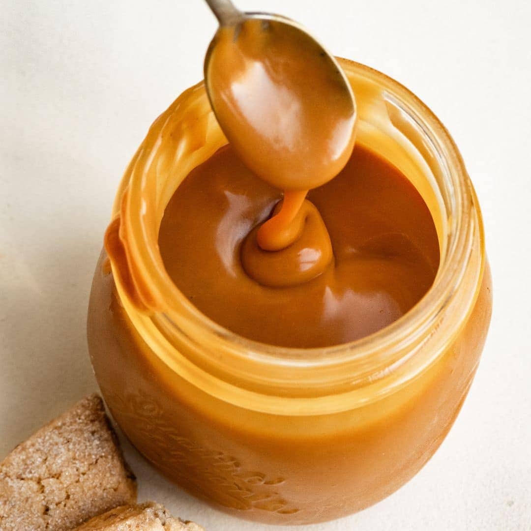 Glass mason jar full of caramel, with a spoon drizzling caramel into the jar.
