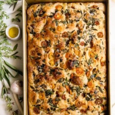 Baked focaccia bread topped with herbs and tomatoes inside a baking pan.