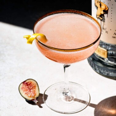 Coupe glass filled with champagne cocktail garnished with a lemon twist next to a sliced fig and bottle of vodka.