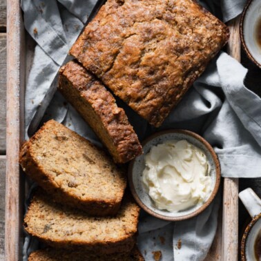 Loaf of banana bread in a wooden tray next to a dish of butter.