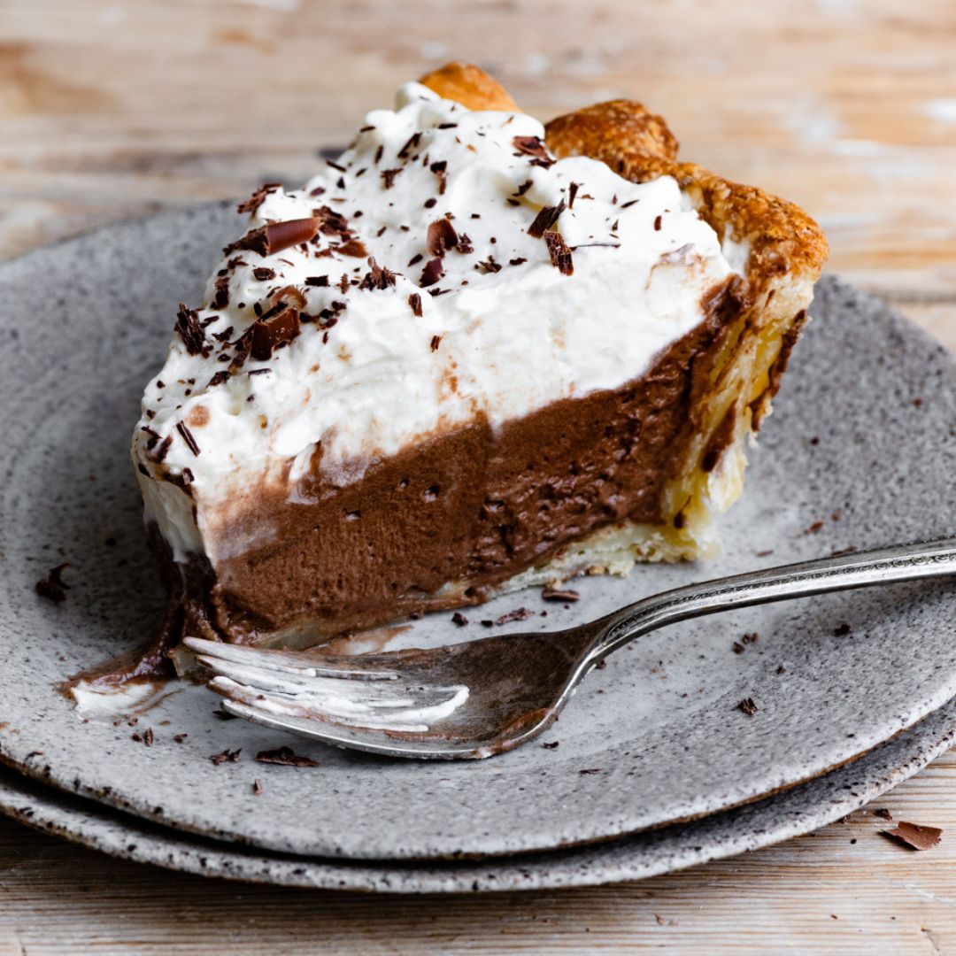 Slice of chocolate pie topped with w hipped cream on a stack of plates next to a fork.