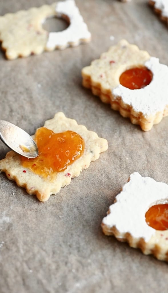 Bottom of a linzer cookie being covered in apricot jam with two sandwich cookies next to it.