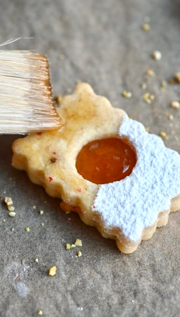 Top of a linzer cookie filled with jam being painted with maple syrup using a pastry brush.