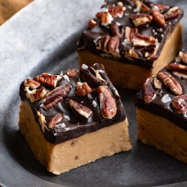 Square pieces of peanut butter fudge topped with chocolate, pecans and salt on a plate.