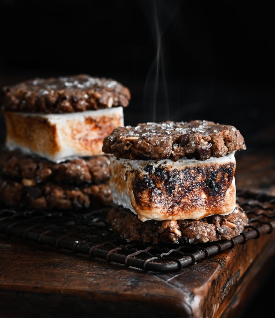 Closeup of two smores made with chocolate chip cookies on a metal tray. The marshmallow filling is toasted and smoking.