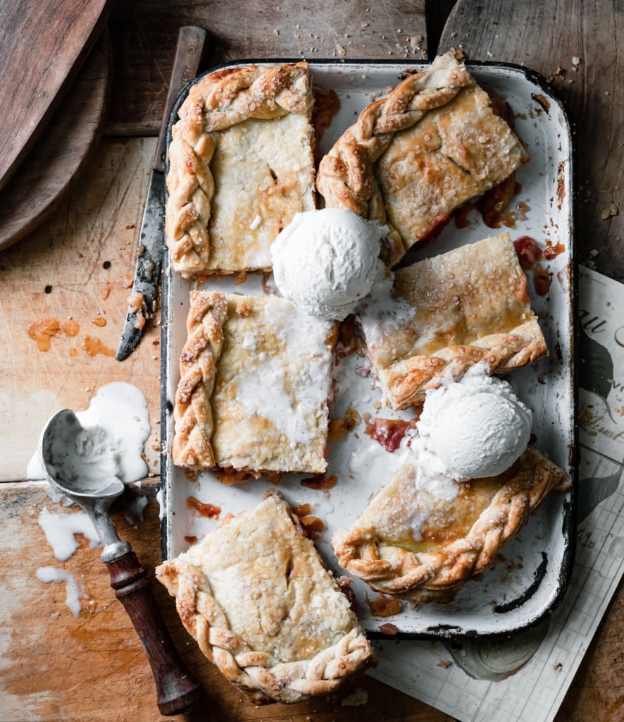 Overhead view of a slab pie on a metal sheet pan topped with scoops of melting vanilla ice cream on a wood table.