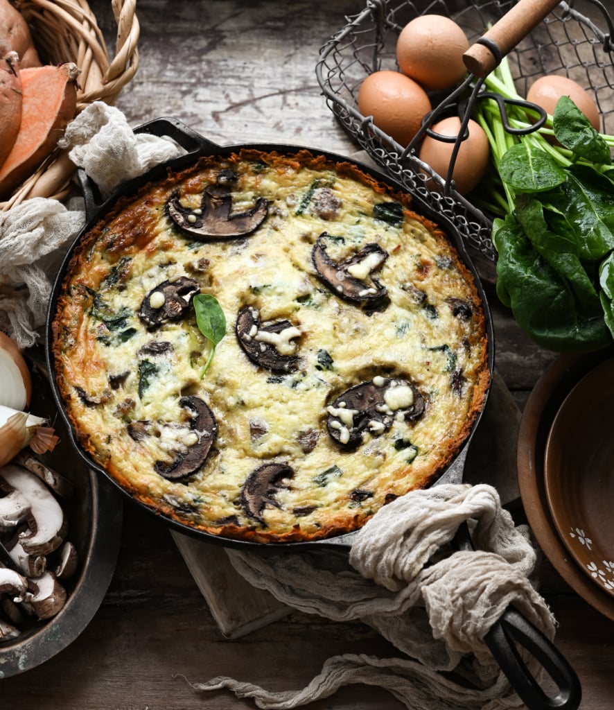 Overhead view of a baked mushroom quiche in a cast iron pan on a wooden table next to a basket of eggs and vegetables.