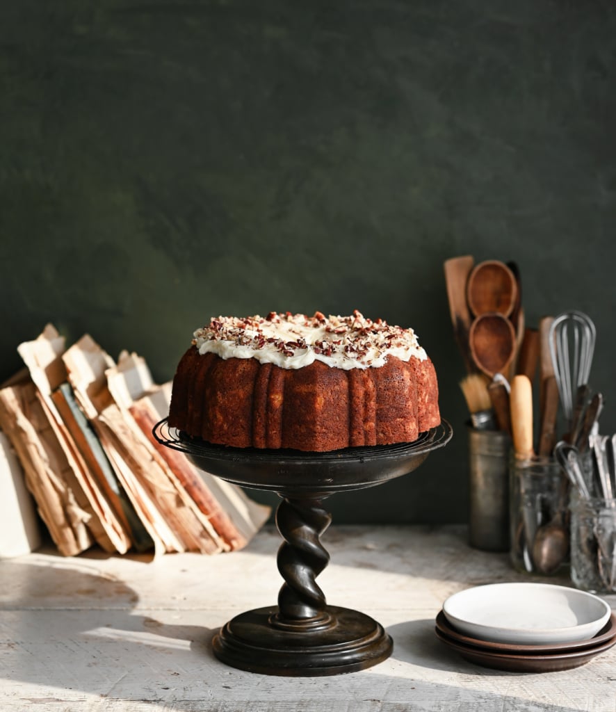 Bundt cake with frosting on a cake stand next to books and jars of baking tools.
