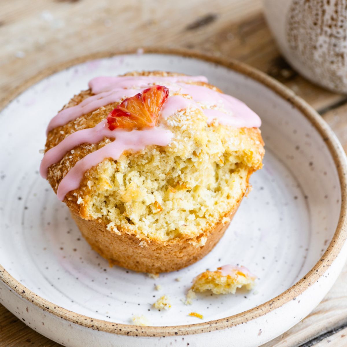 A single orange coconut muffin topped with an orange vanilla glaze and a small orange slice on a plate, with a bite missing.