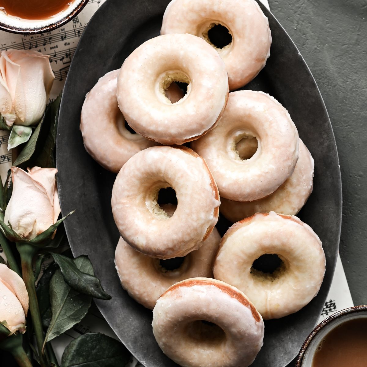 Overhead view of an oval plate stacked full of glazed baked donuts next to two cups of coffee and pink roses.