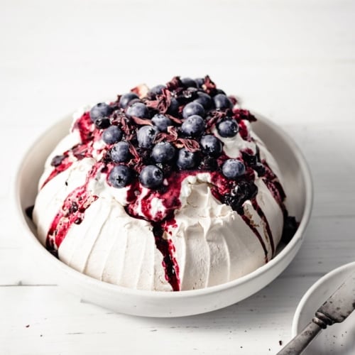 Baked meringue pavlova inside a large serving bowl, topped with whipped cream, hibiscus compote and blueberries, on a wood table.
