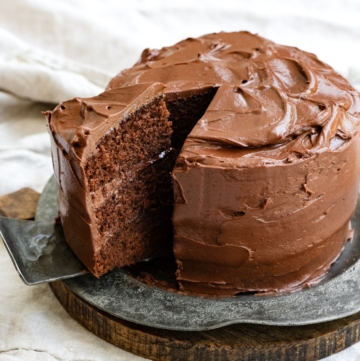 A three layered chocolate cake with chocolate frosting on a metal plate with a slice being pulled out in a pie server.