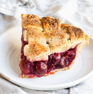 Slice of cherry pie with a lattice crust on a small plate, set on a linen covered table.