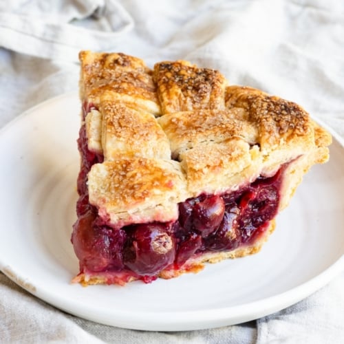 Slice of cherry pie with a lattice crust on a small plate, set on a linen covered table.