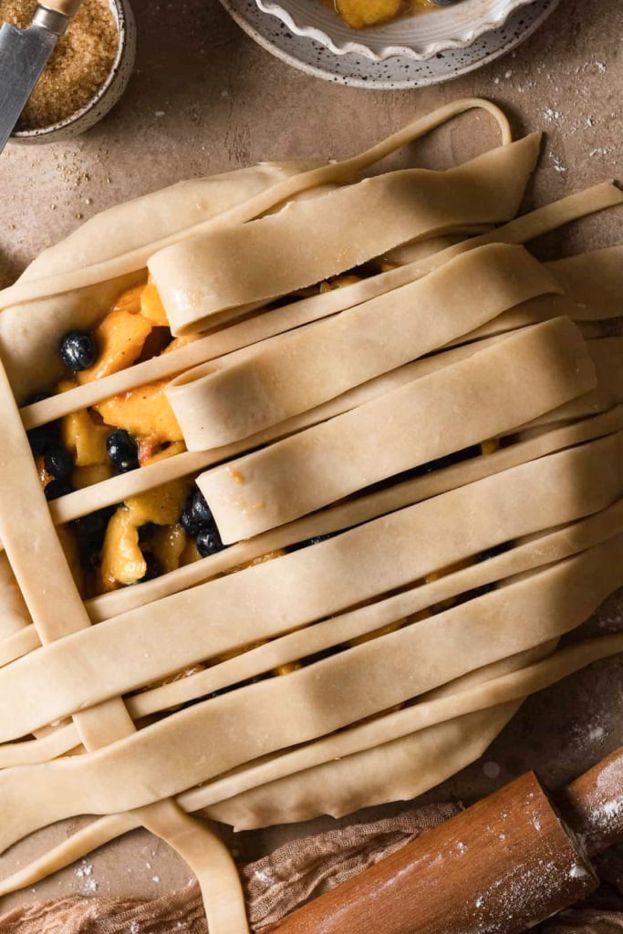 Overhead view of an unbaked pie with strips of lattice pie dough in different thicknesses across the top. Some of the strips are pulled back to show the filling.