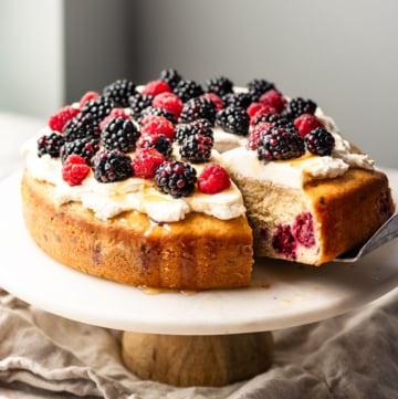 Single layer cake topped with cream and berries on a cake stand with a slice pulled out.