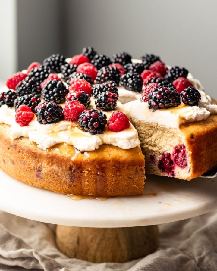 Single layer cake topped with cream and berries on a cake stand with a slice pulled out.