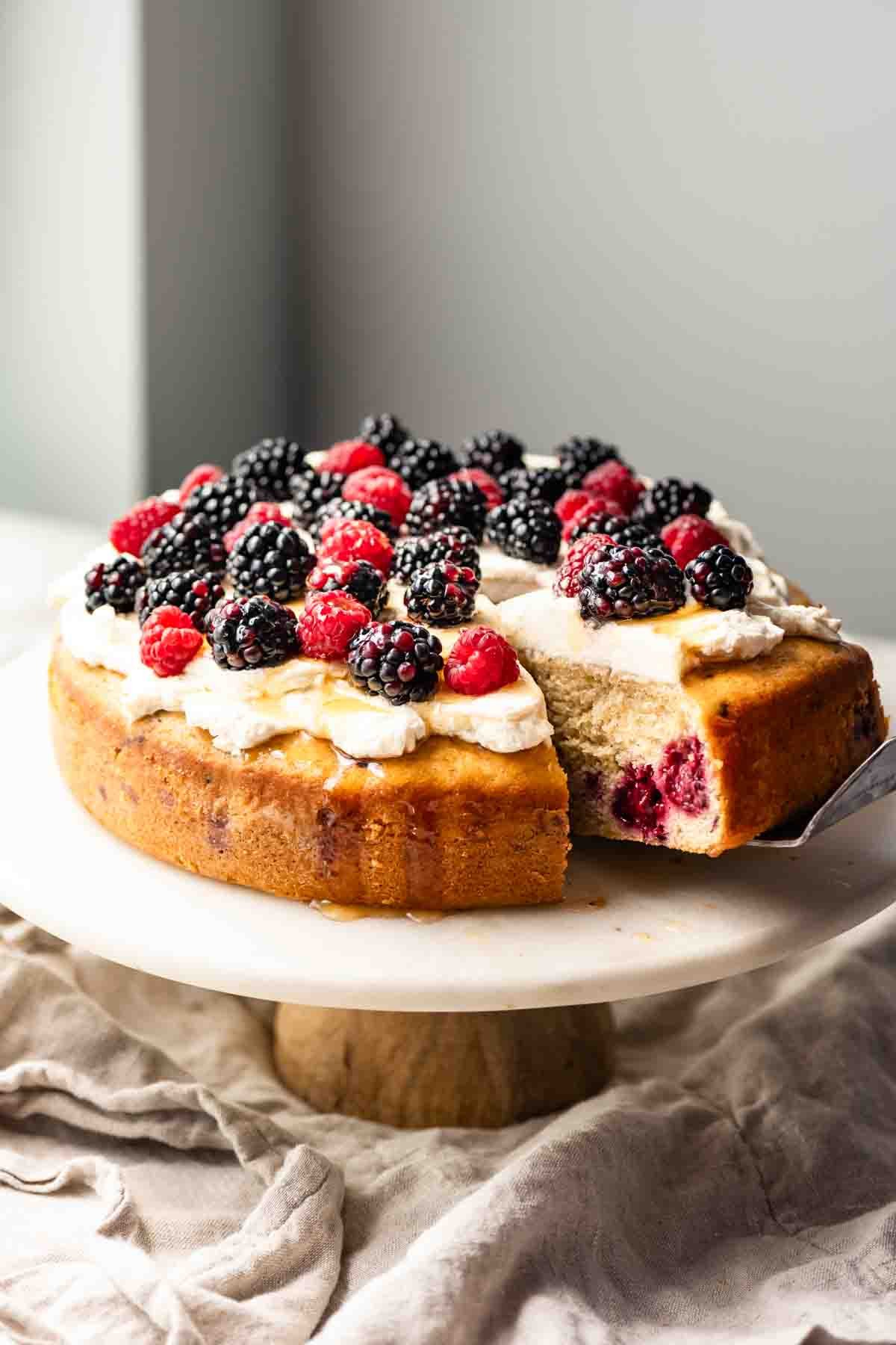 Single layer cake topped with cream and berries on a cake stand with a slice pulled out.