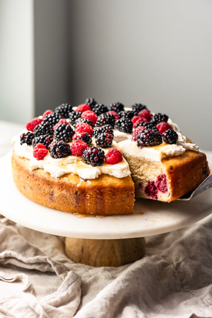 Single layer cake topped with cream and berries on a cake stand with a slice pulled out.