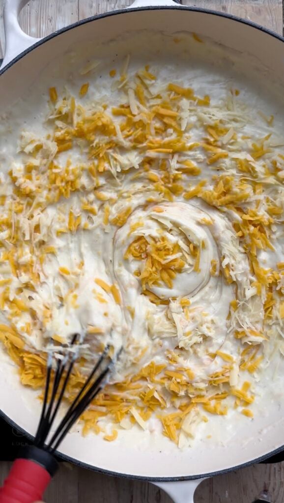 Grated cheese being stirred into a pan full of creamy sauce.
